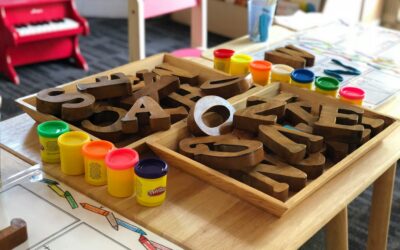 Close-up of a wooden letter set in a tray with colorful Play-Doh containers and a drawing/coloring page on a child's craft table in the background.