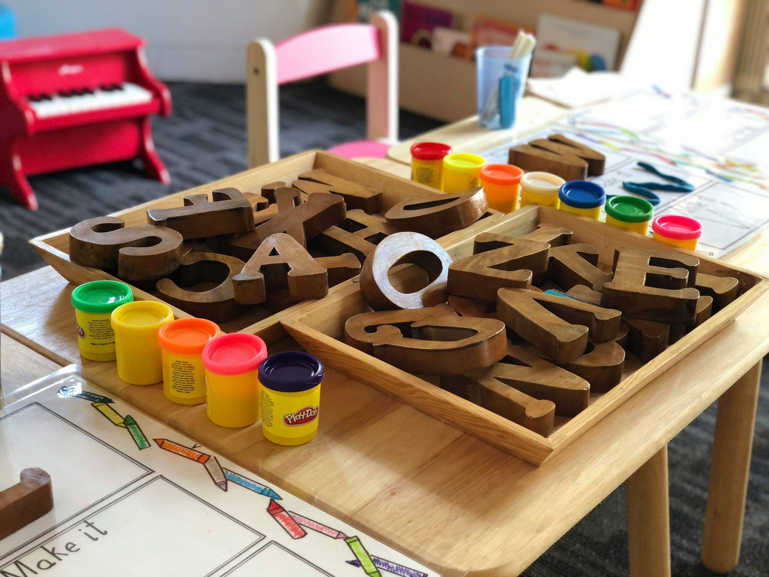 Close-up of a wooden letter set in a tray with colorful Play-Doh containers and a drawing/coloring page on a child's craft table in the background.