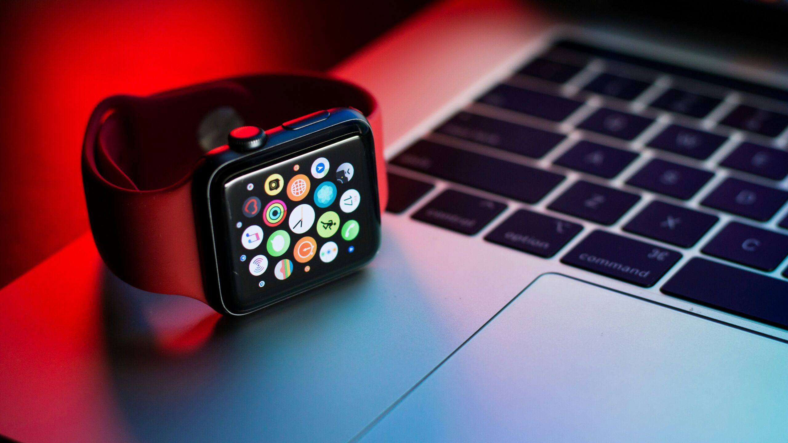 Apple Watch with colorful app icons resting on a laptop keyboard under red and blue lighting