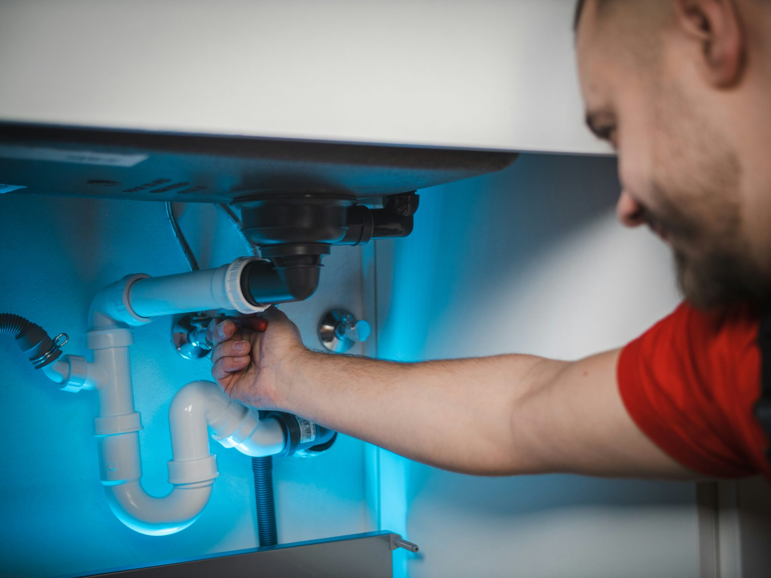 A person tightening plumbing under a sink, with visible PVC pipes and blue-tinted lighting.