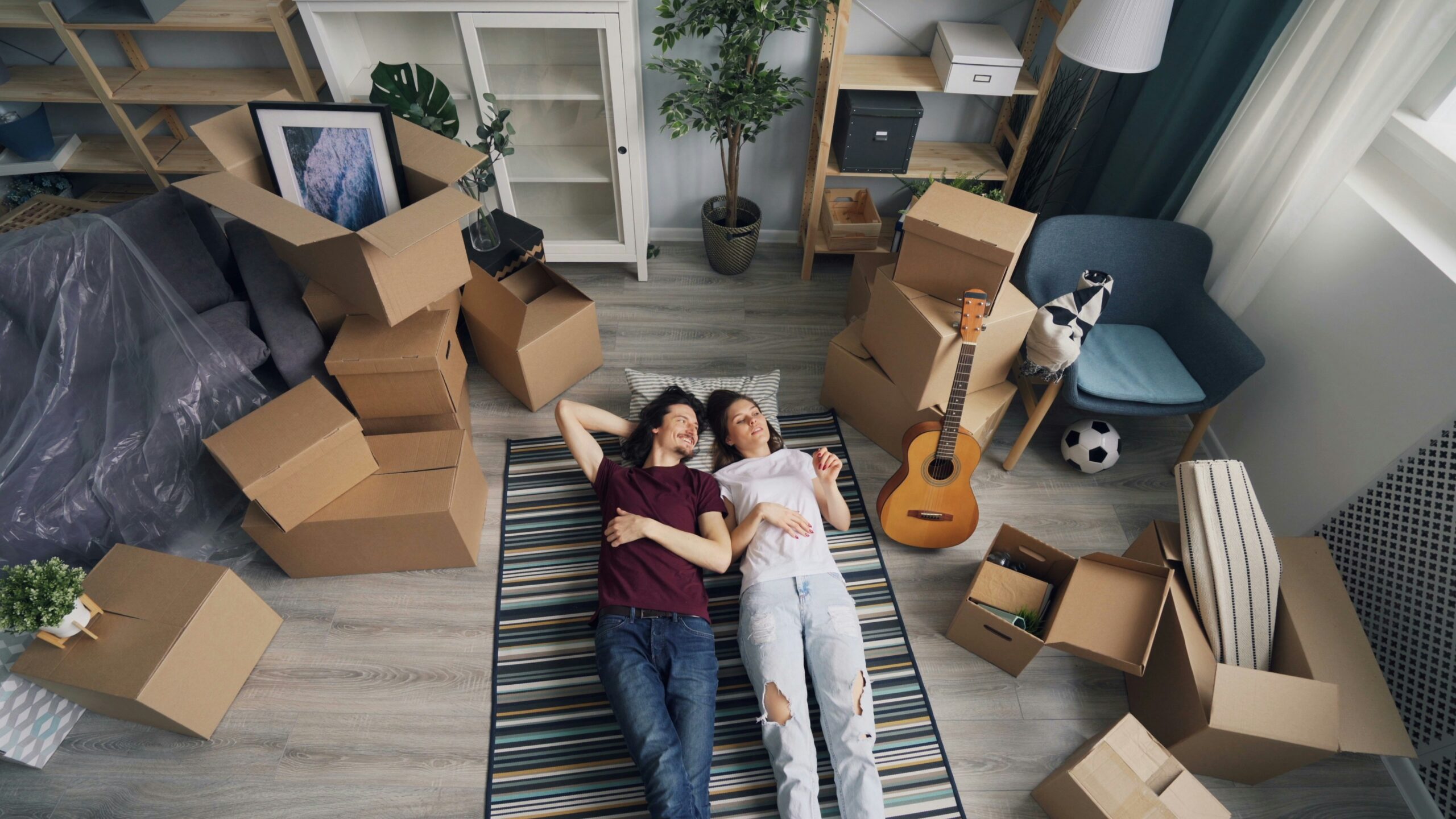 A couple lying on a striped rug on the floor, smiling, surrounded by open and closed moving boxes in a cluttered living room with a guitar nearby.