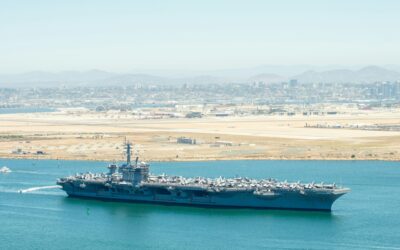 Aircraft carrier with a full flight deck of planes sailing in a blue-green harbor, city and mountains in the distance.