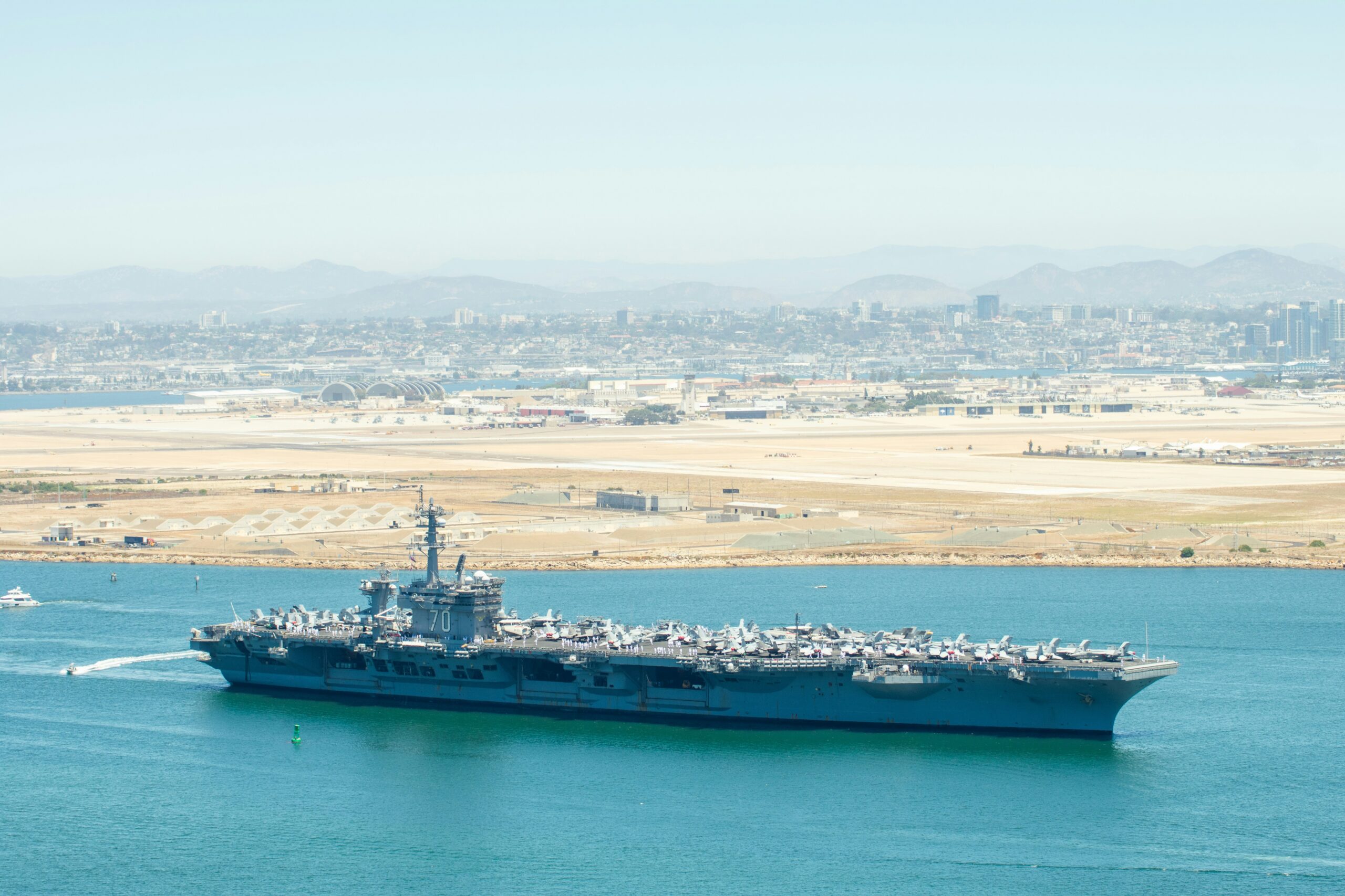 Aircraft carrier with a full flight deck of planes sailing in a blue-green harbor, city and mountains in the distance.