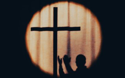 Backlit cross silhouette inside a glowing circular halo against a dark background, with small silhouettes of people in the foreground.