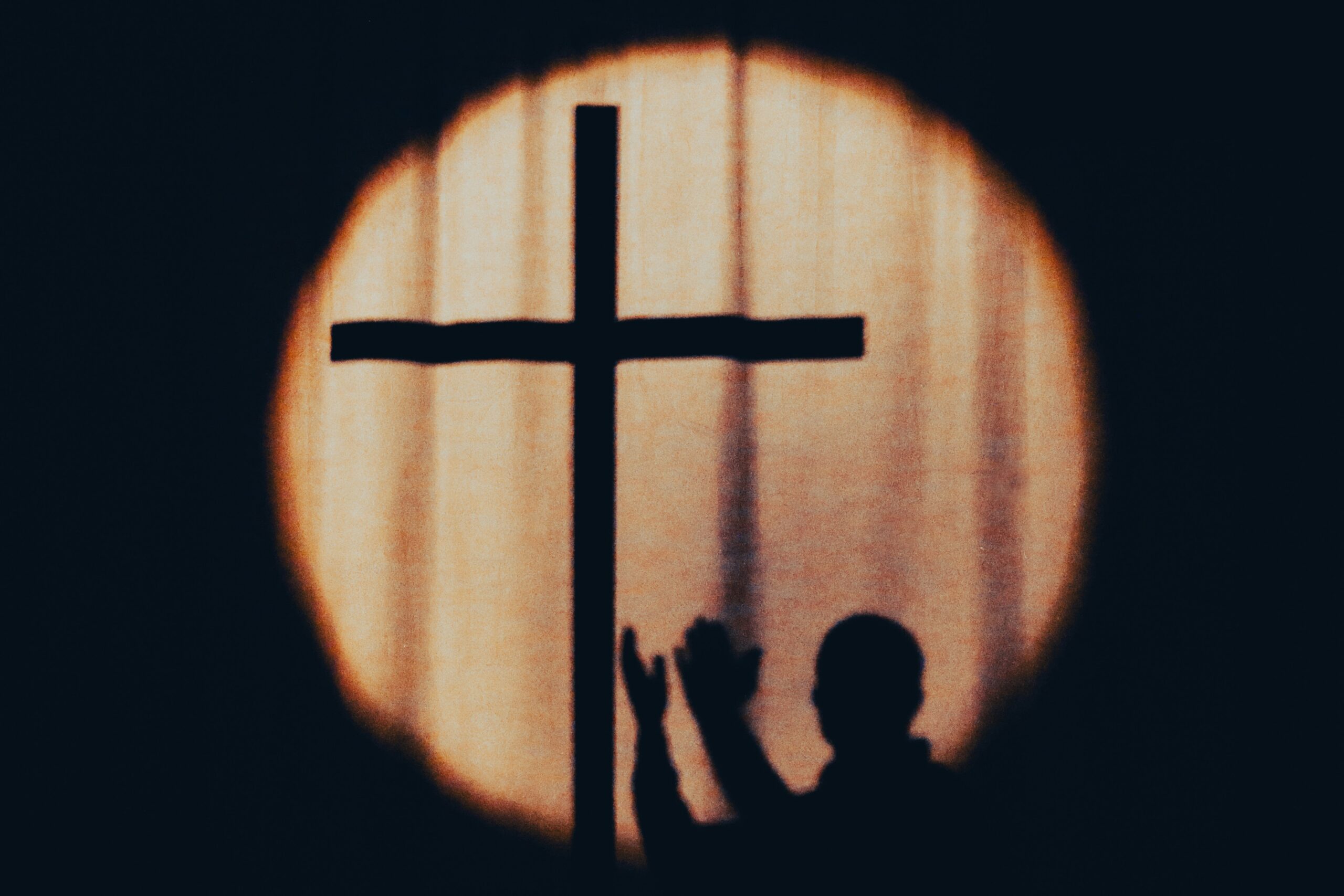 Backlit cross silhouette inside a glowing circular halo against a dark background, with small silhouettes of people in the foreground.
