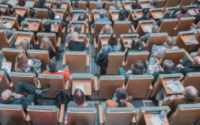 Overhead view of a large lecture hall filled with attendees seated in orange desks, many with papers, tablets, and cups on the desks.