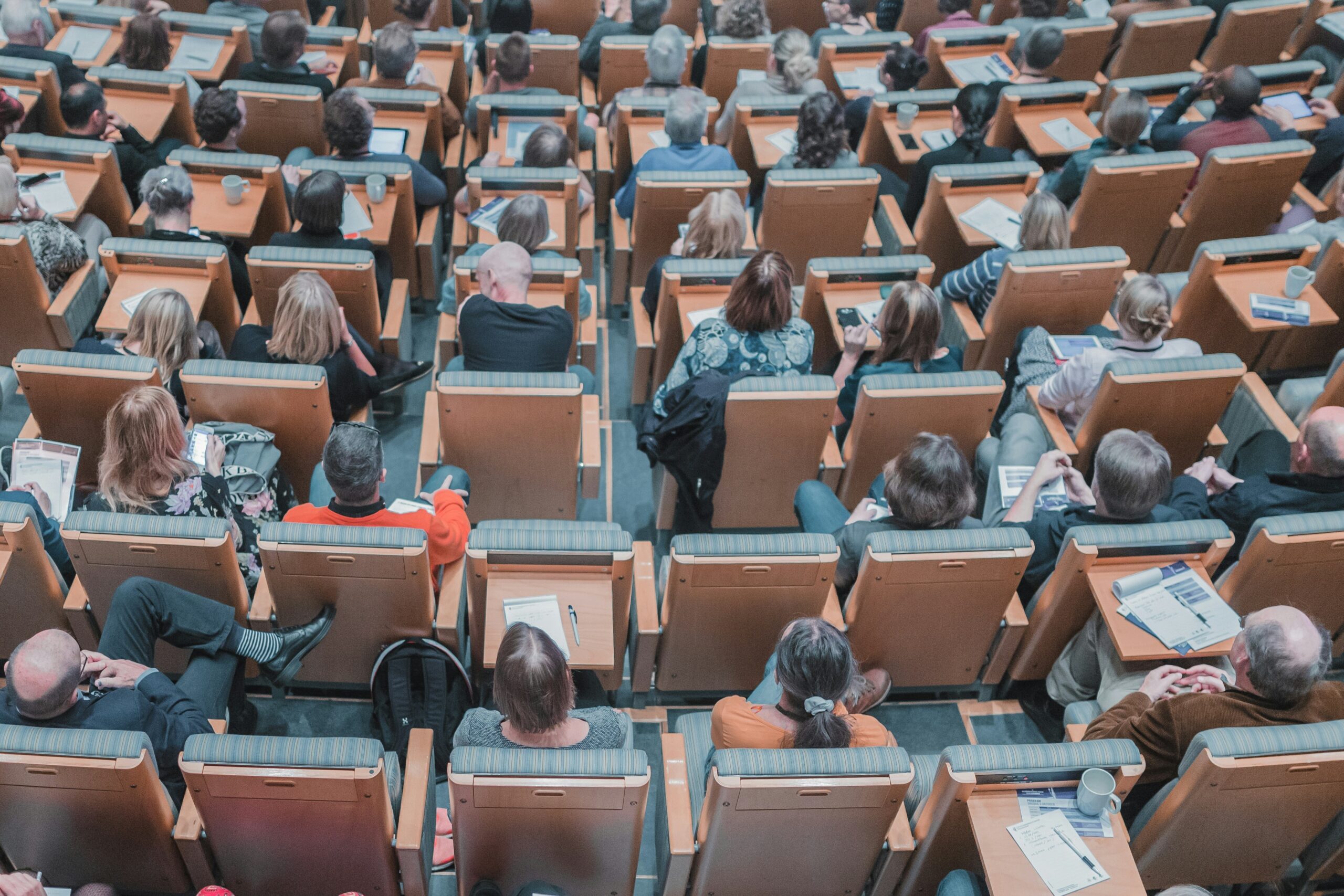 Overhead view of a large lecture hall filled with attendees seated in orange desks, many with papers, tablets, and cups on the desks.