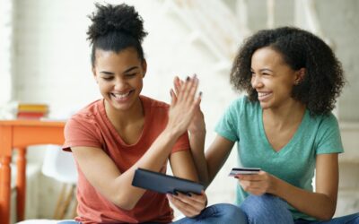 Two young women smile and high‑five while sitting indoors; one holds a tablet, the other a card/card-sized item, showing a moment of celebration or collaboration.