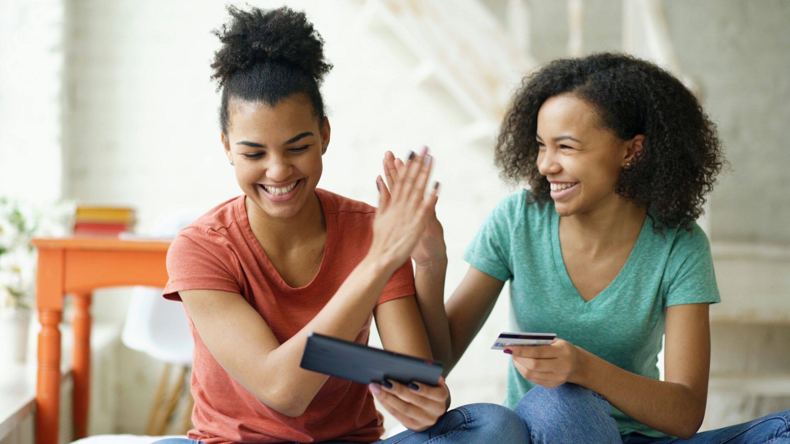 Two young women smile and high‑five while sitting indoors; one holds a tablet, the other a card/card-sized item, showing a moment of celebration or collaboration.