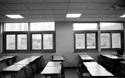 Black-and-white classroom with rows of desks and chairs facing large windows overlooking a cityscape. External light fills the room.