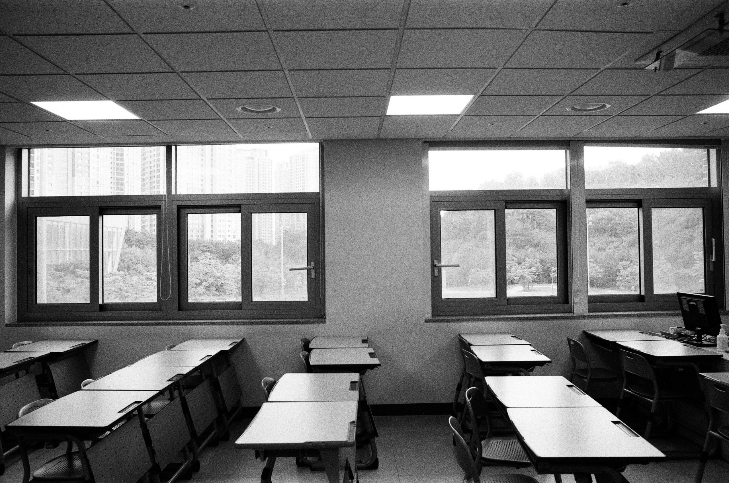 Black-and-white classroom with rows of desks and chairs facing large windows overlooking a cityscape. External light fills the room.