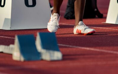 Athlete's white running shoes with orange stripes at the starting blocks on a red track.