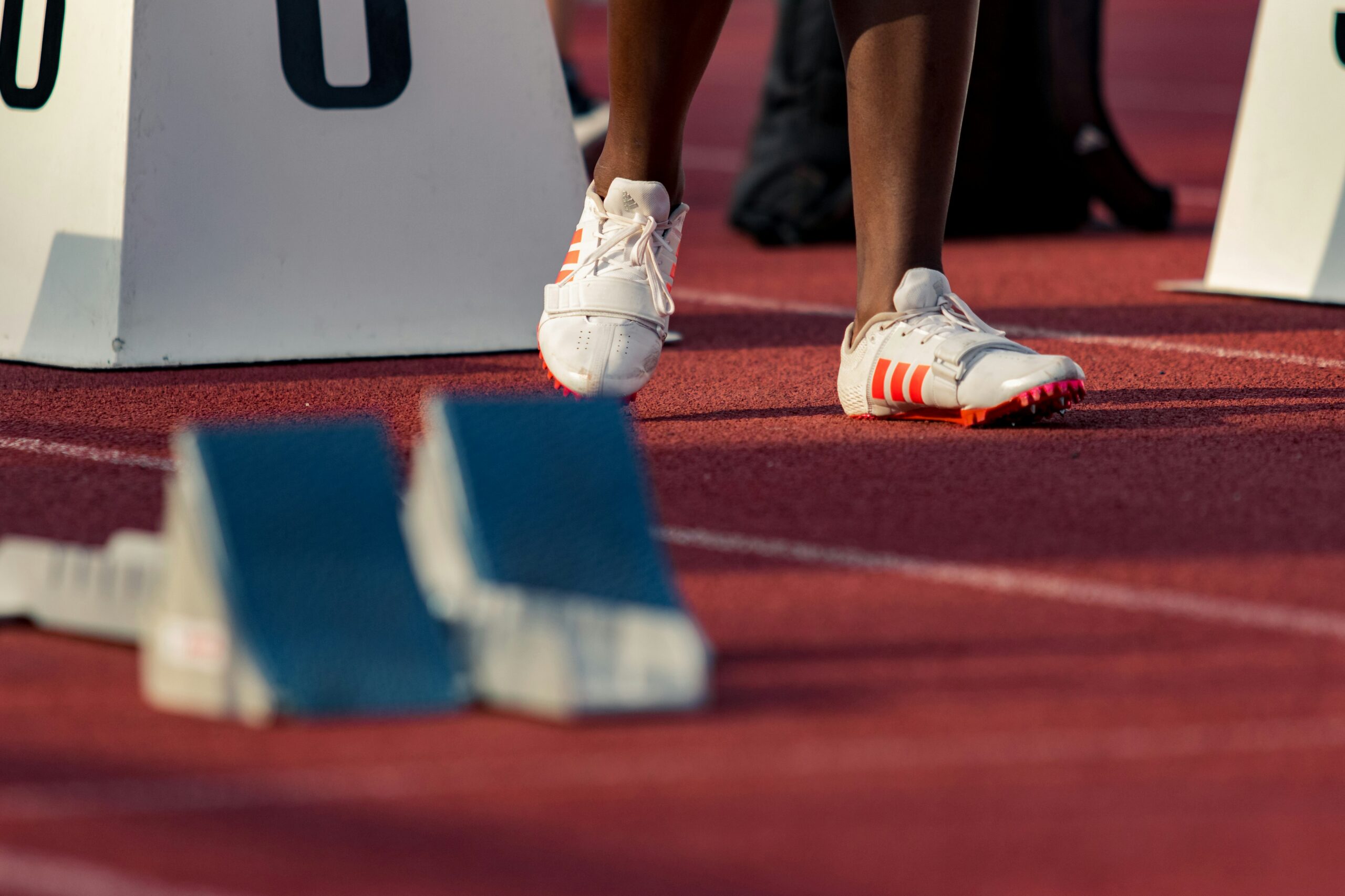 Athlete's white running shoes with orange stripes at the starting blocks on a red track.