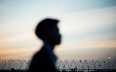 Blurred silhouette of a person walking past a barbed-wire fence at sunset.