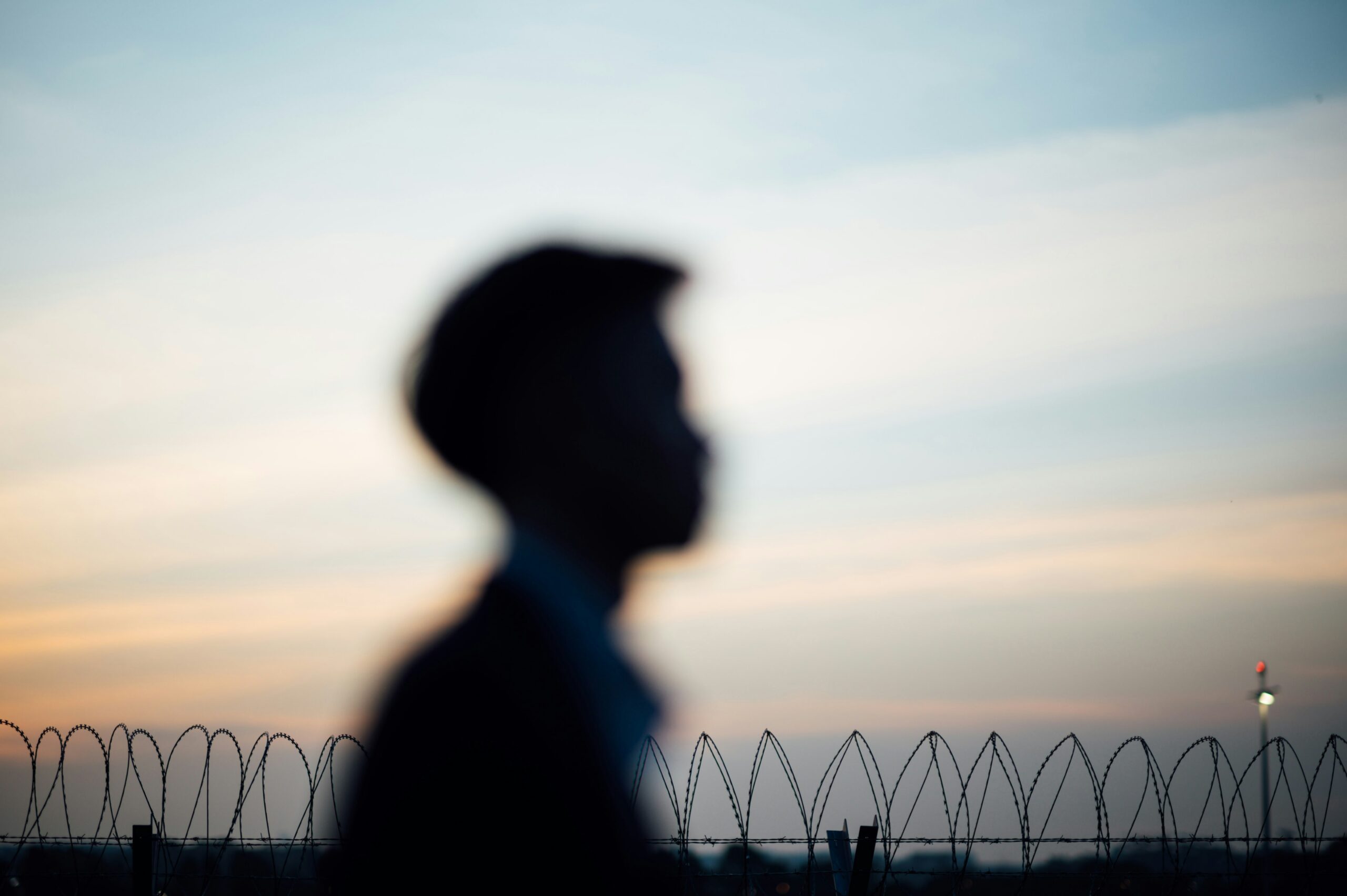 Blurred silhouette of a person walking past a barbed-wire fence at sunset.