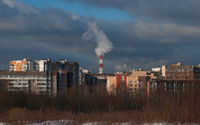 City skyline with apartment buildings and a tall smokestack releasing white smoke under a cloudy sky.