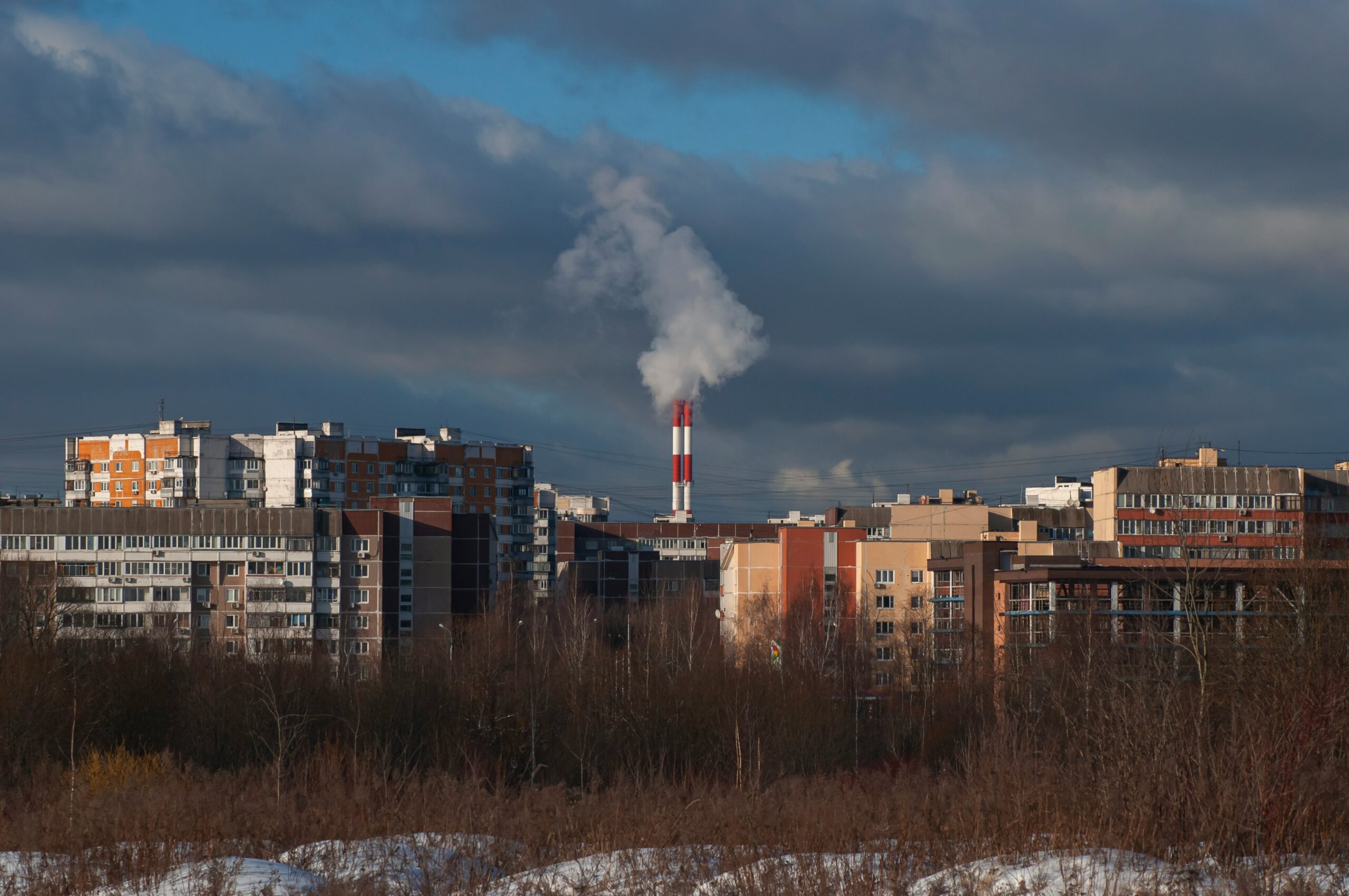 City skyline with apartment buildings and a tall smokestack releasing white smoke under a cloudy sky.