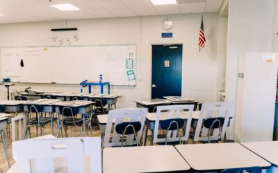 Empty classroom with rows of desks and blue chairs facing a whiteboard and a blue door; American flag and clock on the wall.