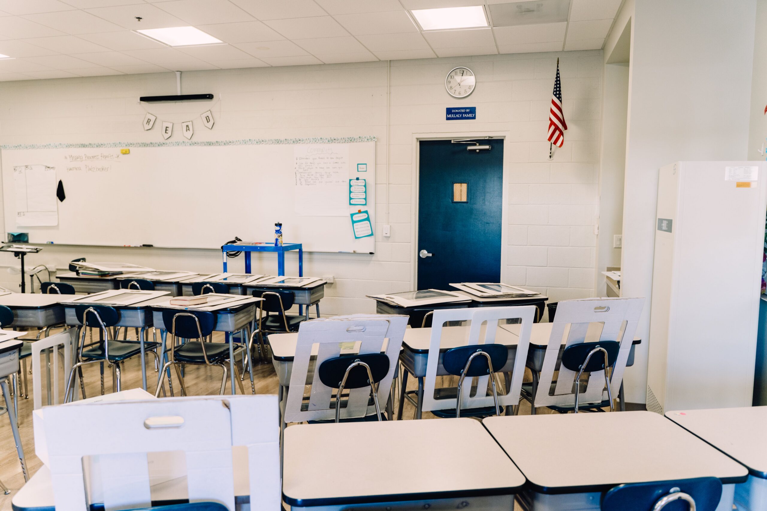 Empty classroom with rows of desks and blue chairs facing a whiteboard and a blue door; American flag and clock on the wall.
