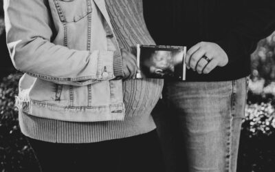 Couple standing close, one in a denim jacket, holding an ultrasound scan of a fetus between them.