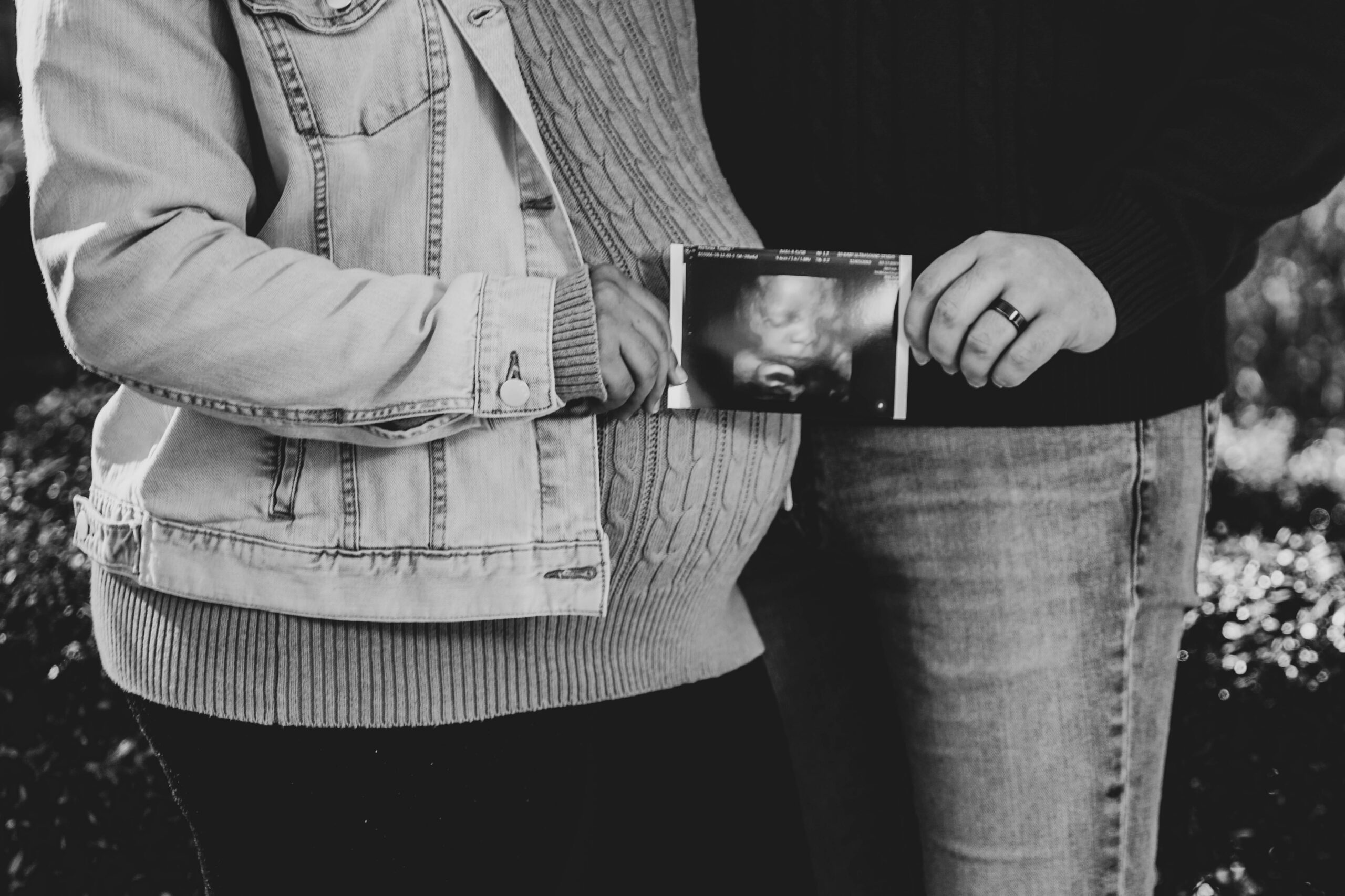 Couple standing close, one in a denim jacket, holding an ultrasound scan of a fetus between them.