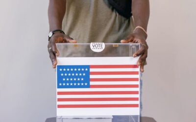 Person placing a ballot into a transparent ballot box with an American flag design on the ballot image. (informative)