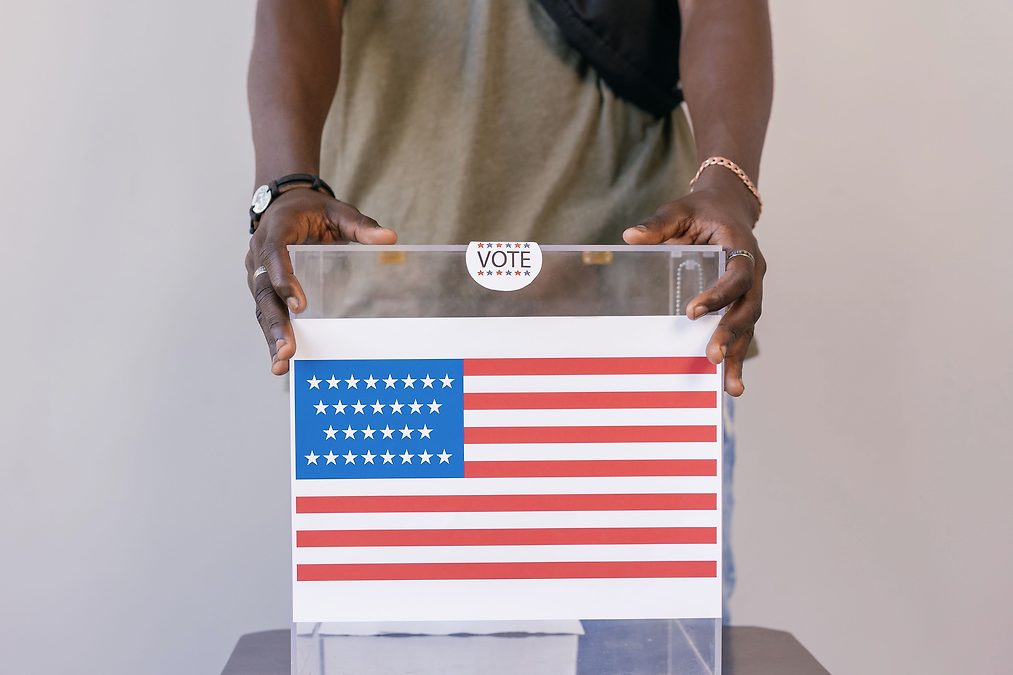 Person placing a ballot into a transparent ballot box with an American flag design on the ballot image. (informative)