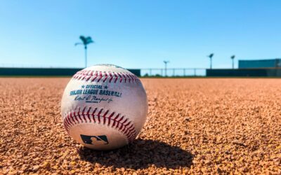 Close-up of a baseball resting on the infield dirt with a clear blue sky overhead.