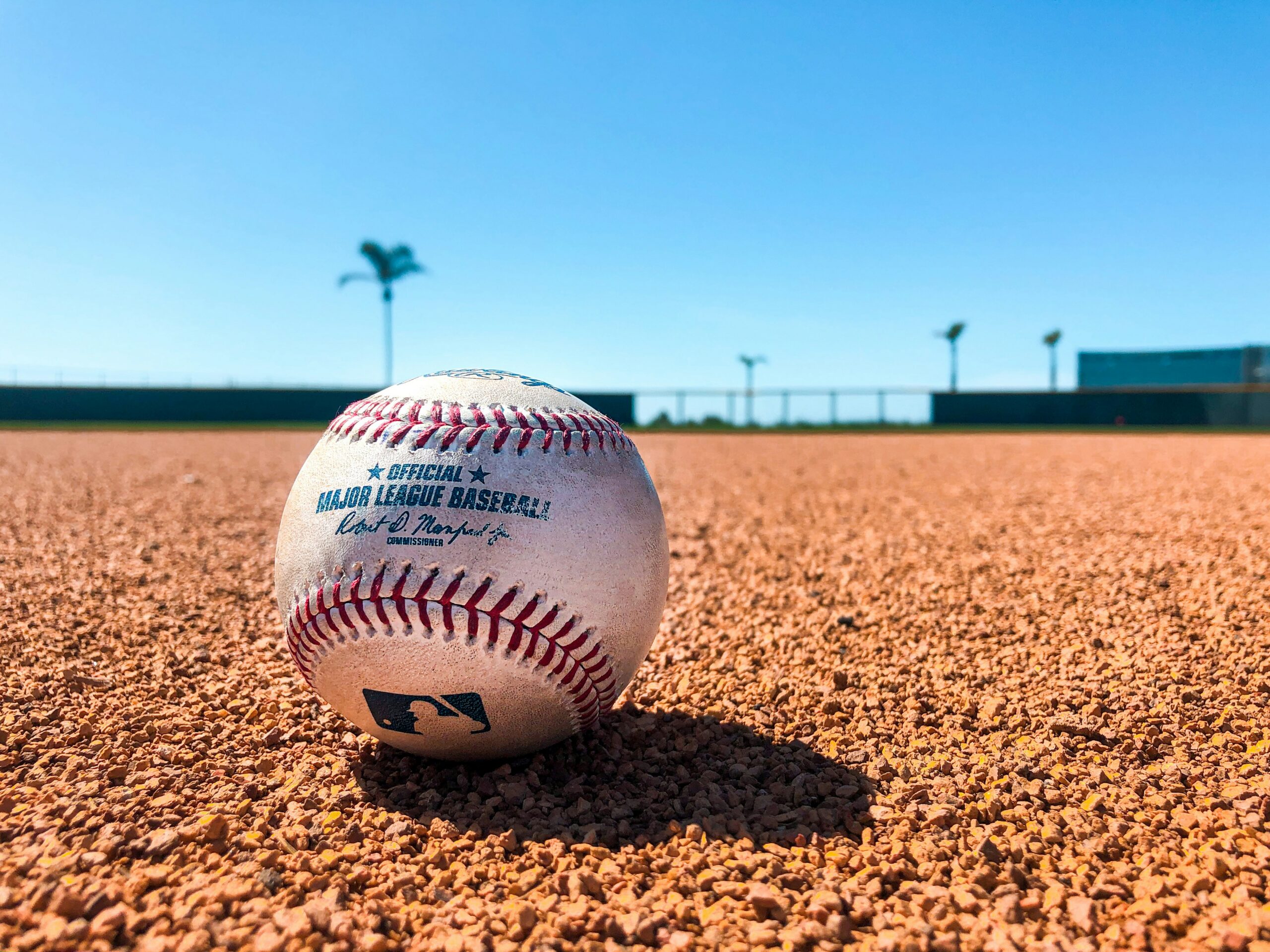 Close-up of a baseball resting on the infield dirt with a clear blue sky overhead.