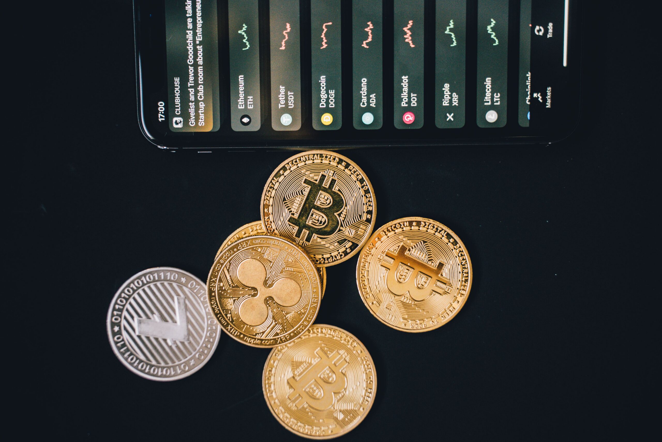 Coins of Bitcoin and other cryptocurrencies on a dark surface with a tilted smartphone displaying a crypto app above them.