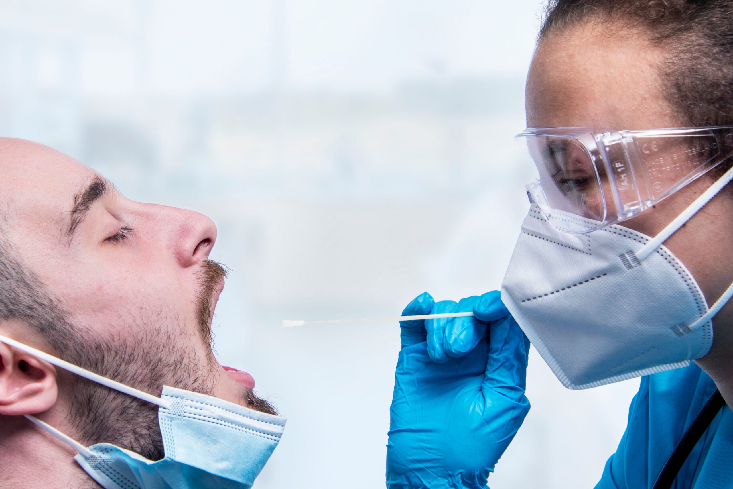 Healthcare worker in goggles and blue gloves swabs a patient’s nose with a cotton swab during a test.
