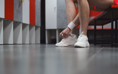 Person in a locker room tying white athletic shoes while seated at a red-and-gray locker row bench.