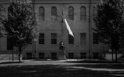 Monument in front of a stone building with a flag on a pole and trees on both sides (black and white photo).