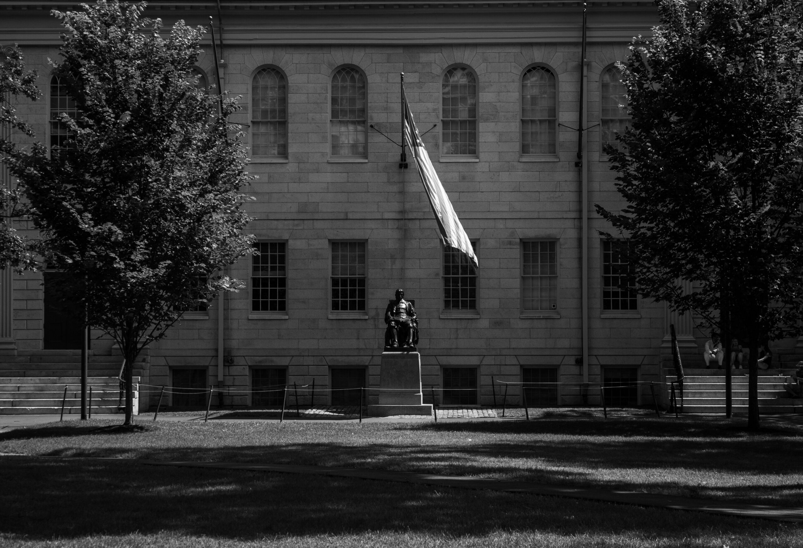 Monument in front of a stone building with a flag on a pole and trees on both sides (black and white photo).