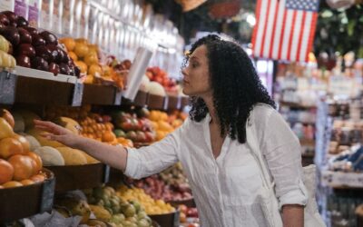 Woman in a white shirt selecting fruit from a grocery store fruit stand, with colorful produce on display and American flag in the background.