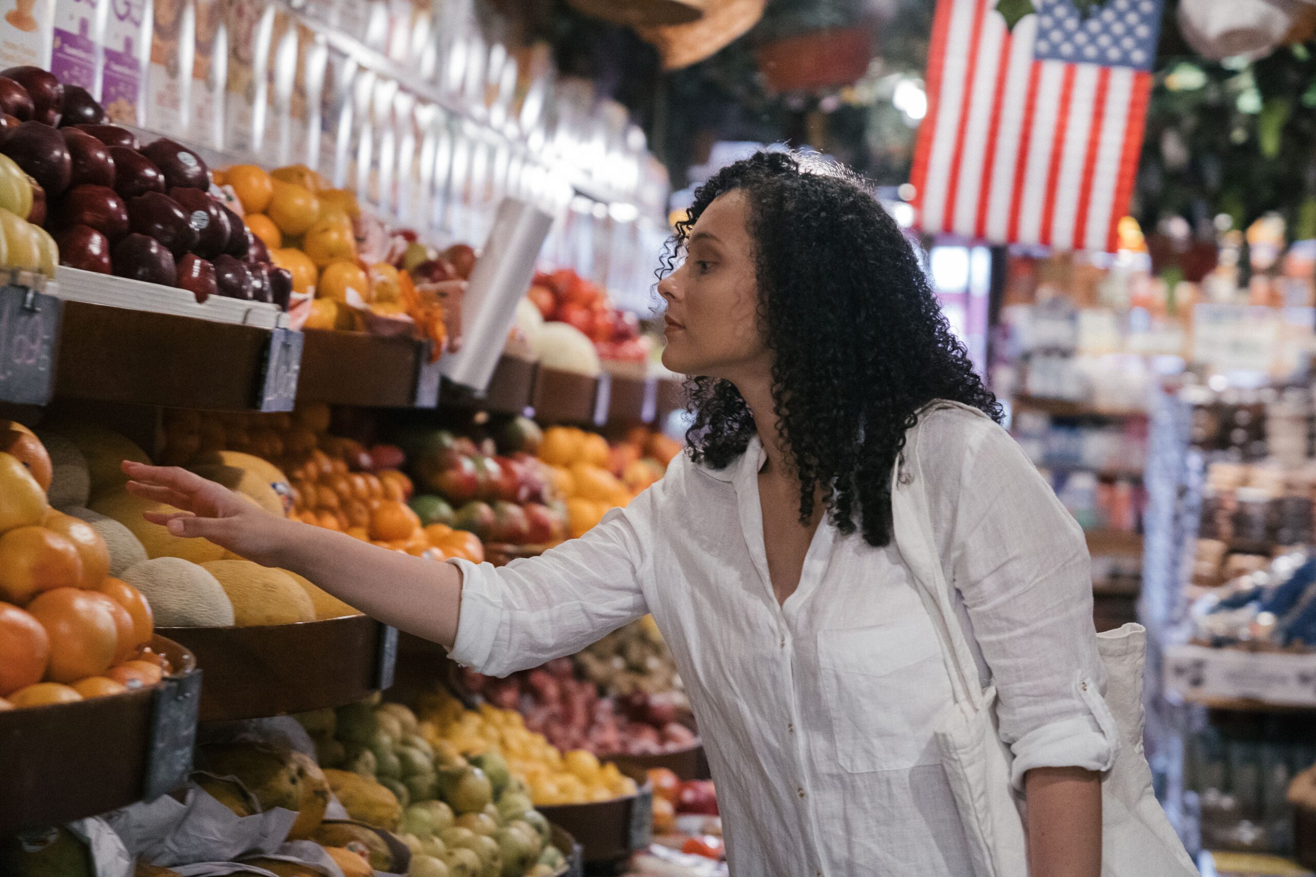 Woman in a white shirt selecting fruit from a grocery store fruit stand, with colorful produce on display and American flag in the background.