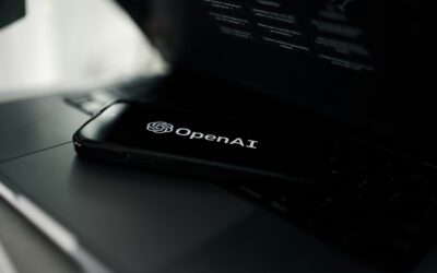 Close-up of a laptop with the OpenAI logo visible on the bottom edge of the screen in a dim setting.
