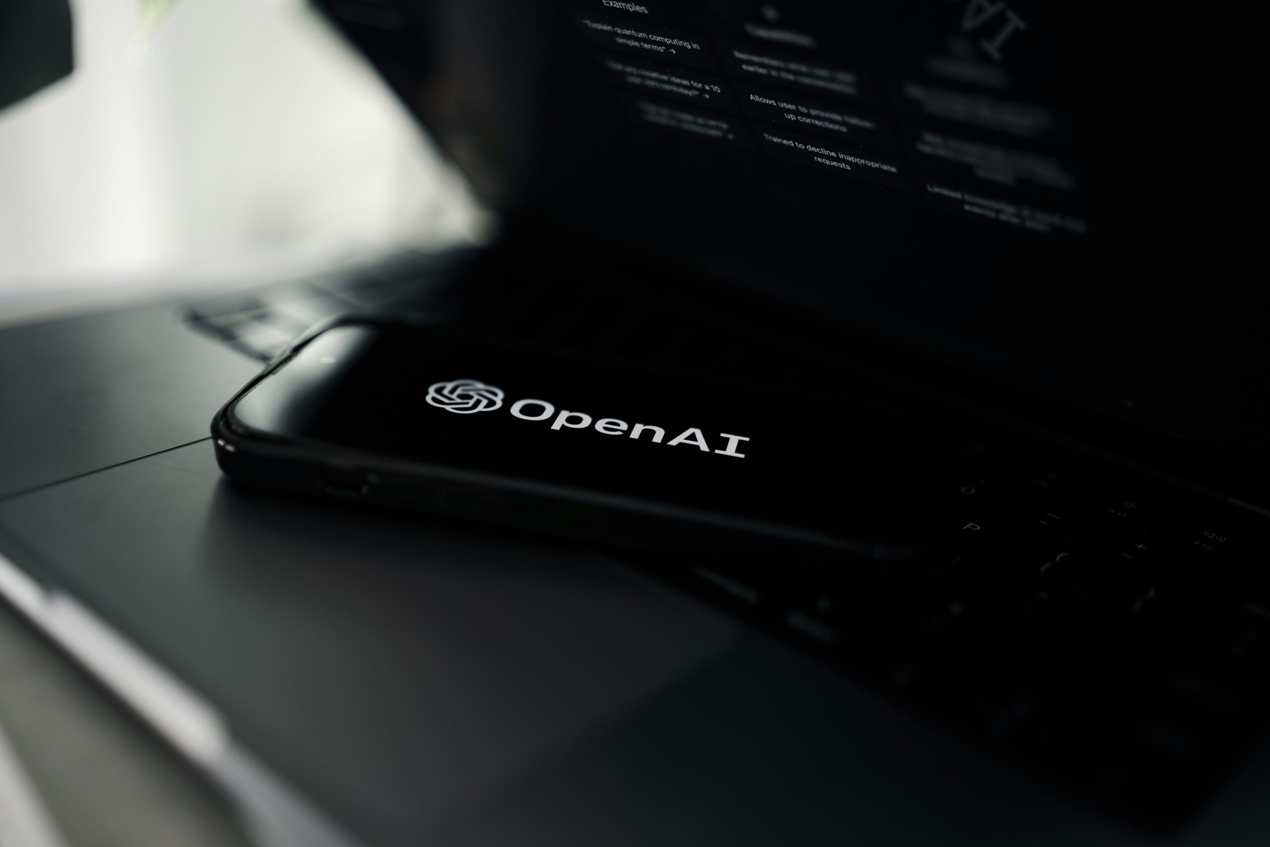 Close-up of a laptop with the OpenAI logo visible on the bottom edge of the screen in a dim setting.